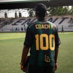 A soccer coach watches a practice session on a sunny day at an empty stadium.